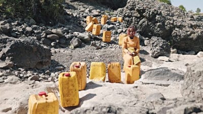 Large yellow water canisters lined up a cliff side, a person wearing yellow is seated amongst them